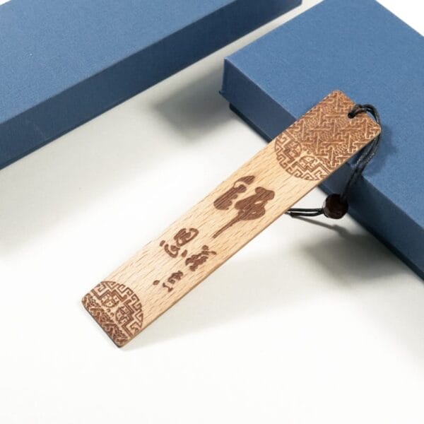 A Chinese style bookmark with intricate carvings and Chinese characters, crafted from redwood engraved bookmarks, is attached to a black string and rests beside two blue boxes on a white surface.