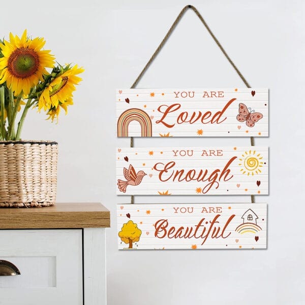 A wooden sign hanging on a wall reads, "You are Loved, You are Enough, You are Beautiful," next to a vase of sunflowers on a white cabinet.