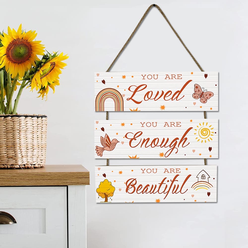 A wooden sign hanging on a wall reads, "You are Loved, You are Enough, You are Beautiful," next to a vase of sunflowers on a white cabinet.