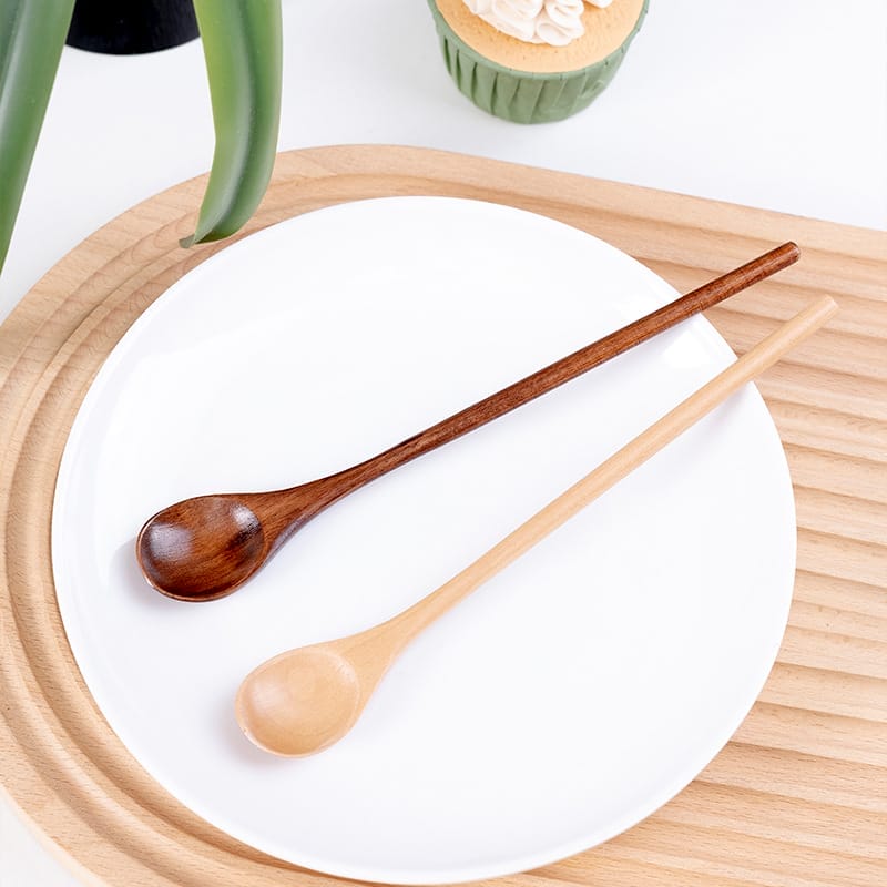Two wooden spoons, one dark and one light, rest on a white plate. Wholesale honey spoons from a wooden spoon manufacturer.
