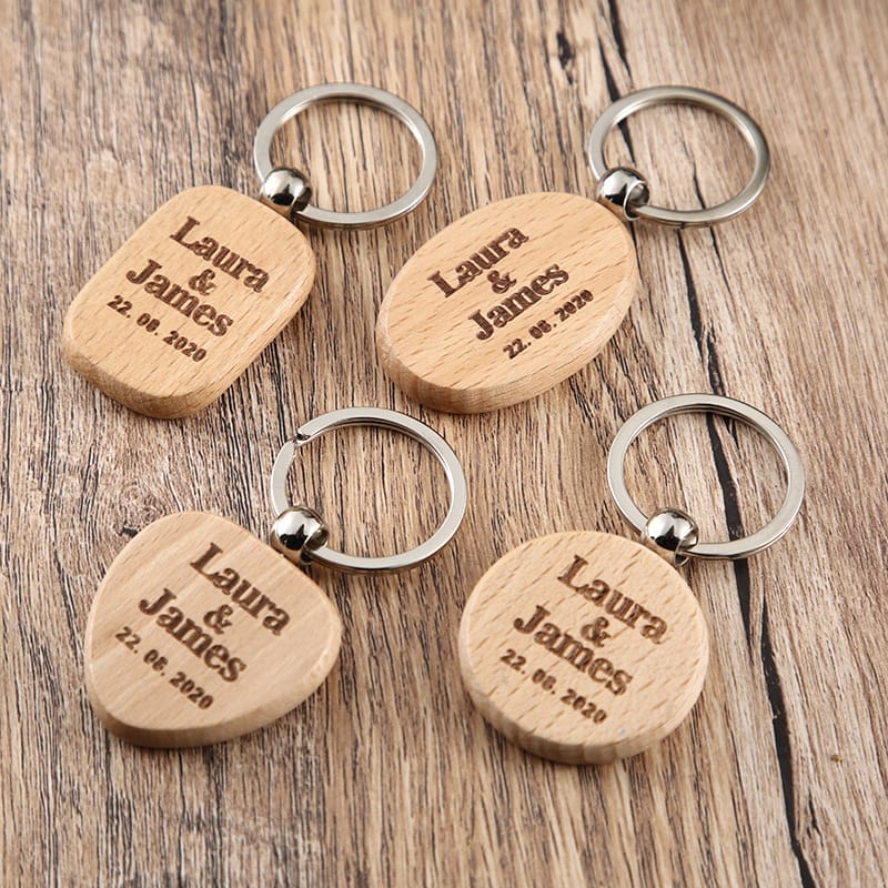 Five keychain wooden pieces engraved with "Laura & James 22.08.2020" are arranged on a wooden surface; each keychain has a unique shape and sturdy metal ring.