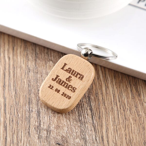 A custom keychain wooden engraved with "Laura & James 22.08.2020" rests on a wooden surface, making it a perfect keepsake.