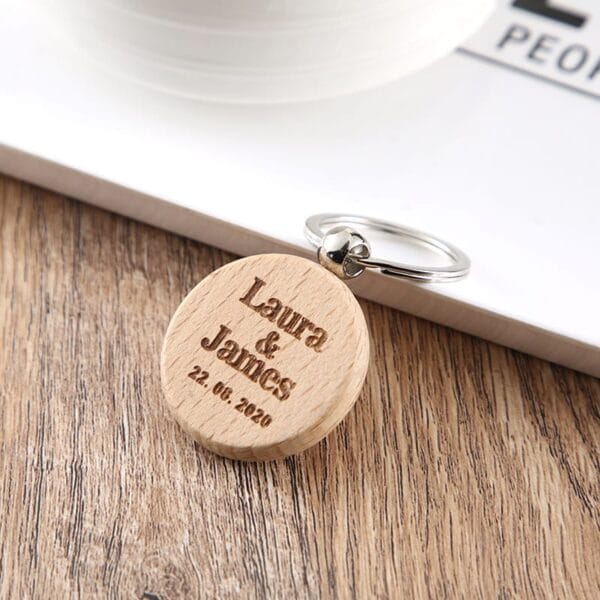 A round custom keychain wooden, engraved with "Laura & James 22.08.2020," sits on a wooden surface next to a book.