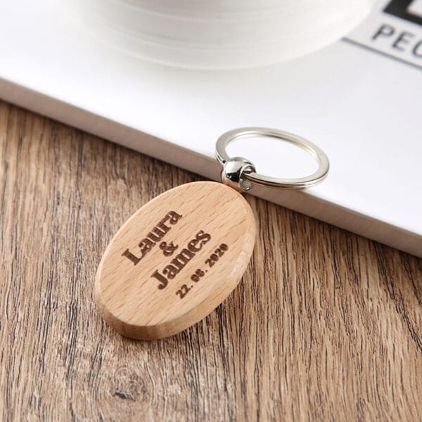 A wooden oval keychain, crafted by a custom keychain manufacturer, is engraved with "Laura & James" and the date "22. 8. 2020," placed on a wooden surface near a white book and cup.