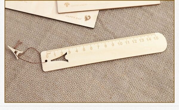 A wooden ruler with centimeter markings from 0 to 15, featuring a cut-out fish shape and a brown string, lies on a textured beige fabric surface—an ideal choice for custom gifts or unique engraved rulers.