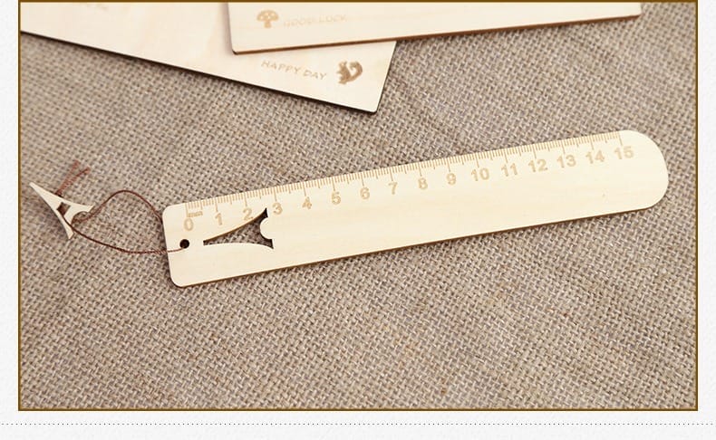 A wooden ruler with centimeter markings from 0 to 15, featuring a cut-out fish shape and a brown string, lies on a textured beige fabric surface—an ideal choice for custom gifts or unique engraved rulers.