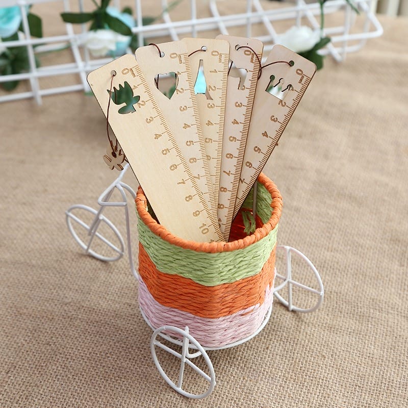 A small white wire tricycle holds a woven basket filled with engraved rulers and wooden laser cut bookmarks, each featuring unique cut-out shapes and designs, standing upright inside—perfect as custom gifts.