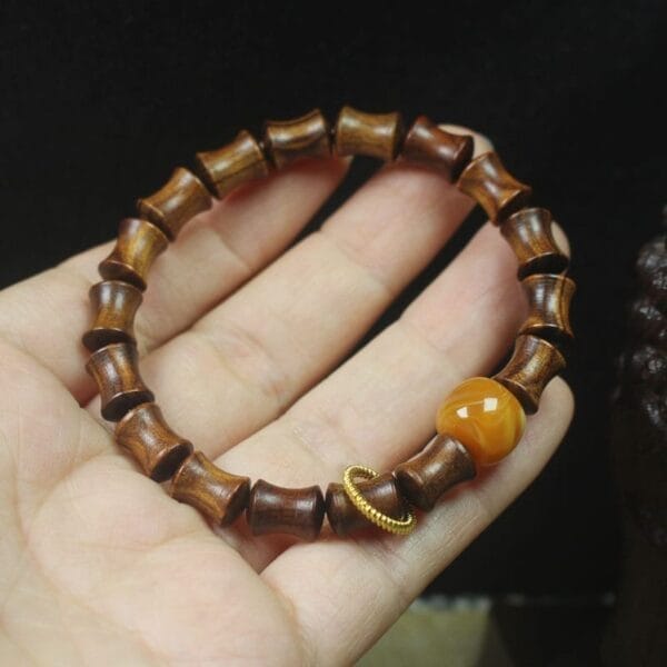 A hand holds a Men’s wood bracelet made of cylindrical wooden beads, featuring one round amber-colored bead and a small gold ring—perfect for those seeking wholesale, unique accessories.