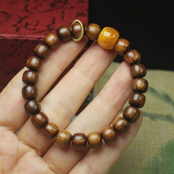 A hand holds a Men’s wood bracelet made of round brown wooden beads, featuring one larger yellow bead and a decorative golden ring.