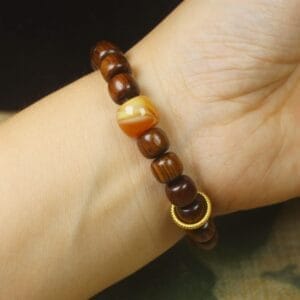 A wrist wearing a men’s wood bracelet featuring dark brown wooden beads, one marbled yellow bead, and two small textured gold rings—an eye-catching piece among durable wristbands.