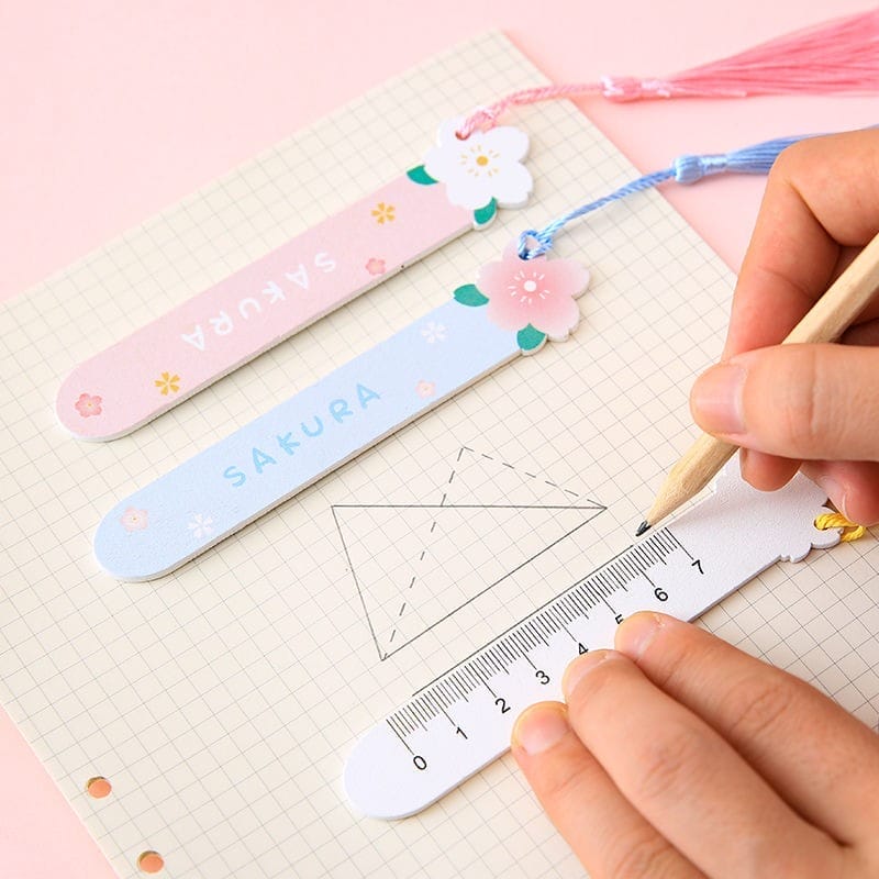 A hand uses a ruler with a flower design to draw on graph paper, with two personalized wooden bookmarks labeled “SAKURA” and adorned with floral accents nearby.