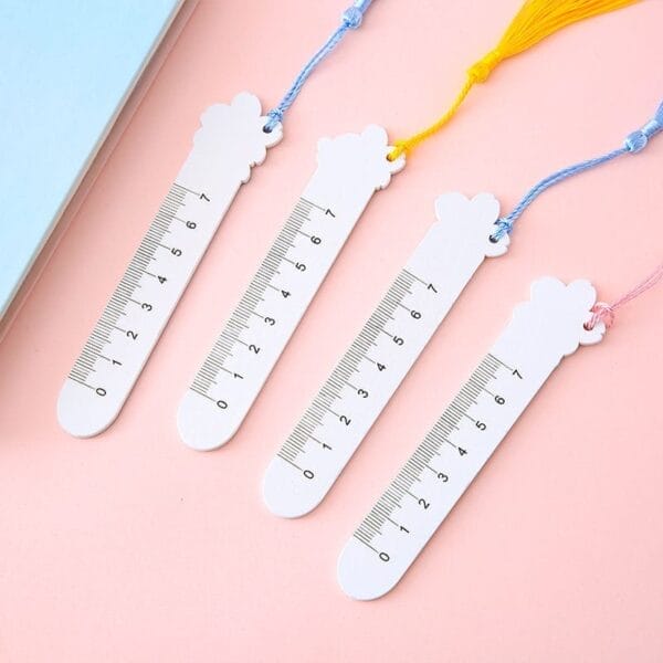 Four white cloud-shaped bookmarks with ruler markings, each attached to a different colored tassel, are laid out on a pink surface—an adorable alternative to personalized wooden bookmarks.