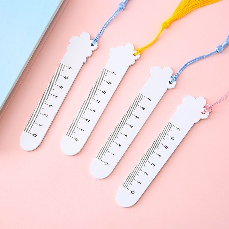 Four white cloud-shaped bookmarks with ruler markings, each attached to a different colored tassel, are laid out on a pink surface—an adorable alternative to personalized wooden bookmarks.