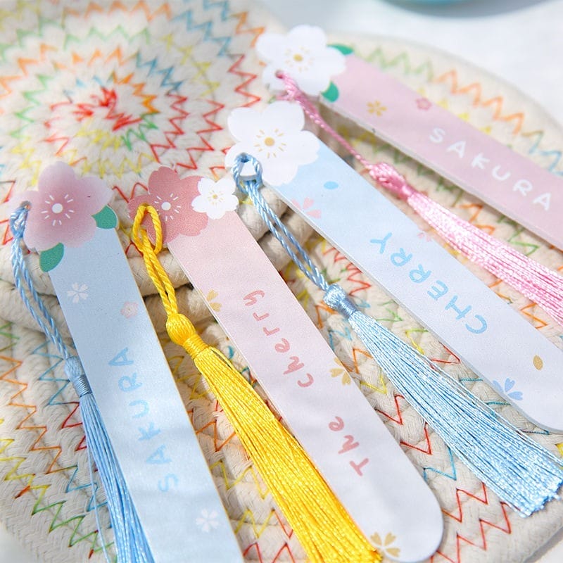 Five pastel-colored, personalized wooden bookmarks with tassels and floral designs, labeled "SAKURA" and "CHERRY," are arranged on a patterned fabric background.