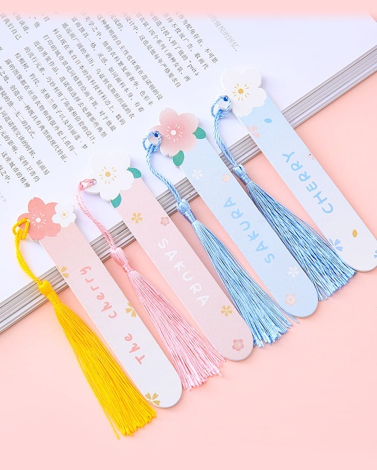 Five pastel-colored personalized wooden bookmarks with floral designs and tassels, labeled "The cherry" and "Sakura," are placed on an open book on a pink surface.