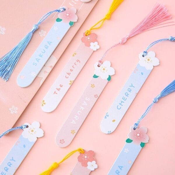 Several pastel-colored bookmarks with floral designs and tassels are arranged on a pink surface. Each personalized wooden bookmark features words like "SAKURA" and "CHERRY.