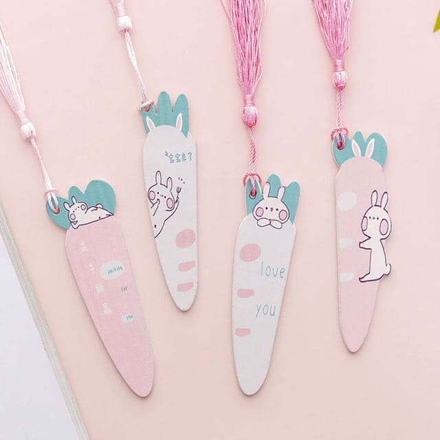Four carrot-shaped personalized wooden bookmarks with pink tassels, featuring cute bunny illustrations and pastel colors, arranged on a light pink surface.