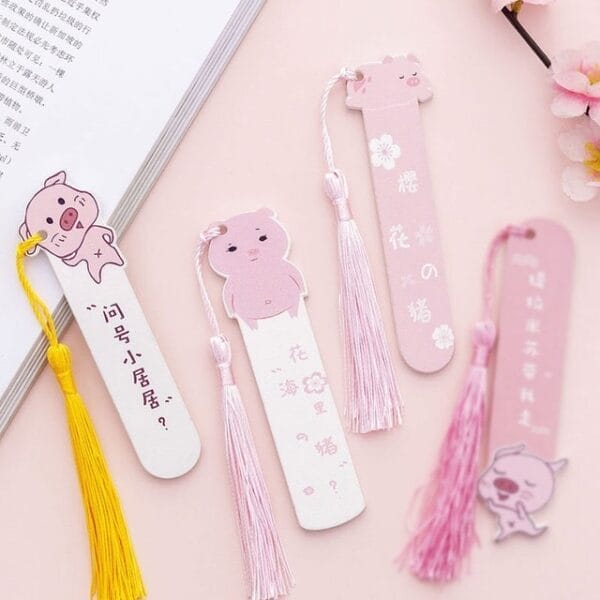 Four pastel-colored Personalized Wooden Bookmarks with pig designs and tassels are arranged on a pink surface, alongside a book and decorative flowers.