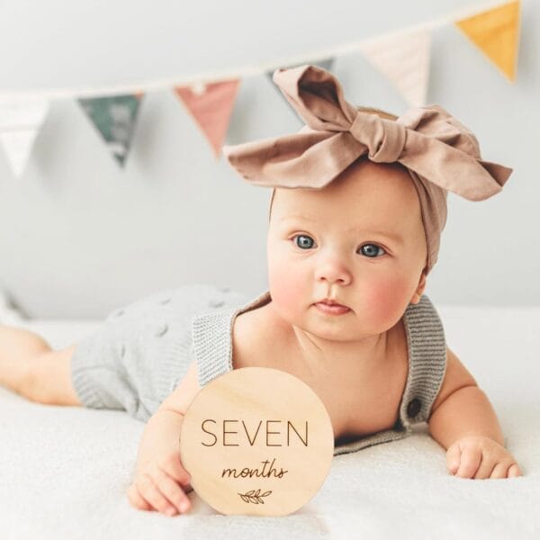A baby wearing a headband and gray outfit lies on a bed, holding a round wood piece that reads "Seven months." Bunting decorates the background.