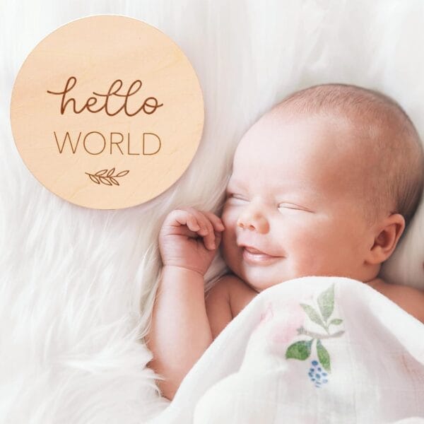 A newborn baby lies on a white fluffy blanket, partially covered with a floral cloth, next to a round wood piece perfect for newborn milestone photos that reads "hello WORLD.