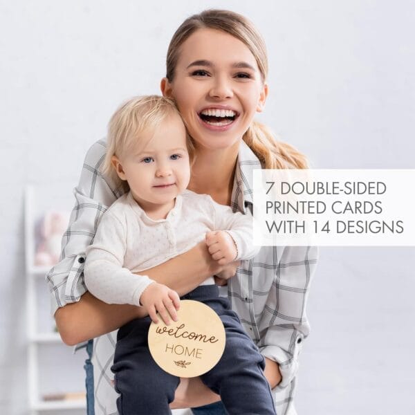 Smiling woman holds a young child who is holding a round wood piece reading "welcome HOME." Text overlay reads "7 double-sided printed cards with 14 designs—perfect for newborn milestone photos.