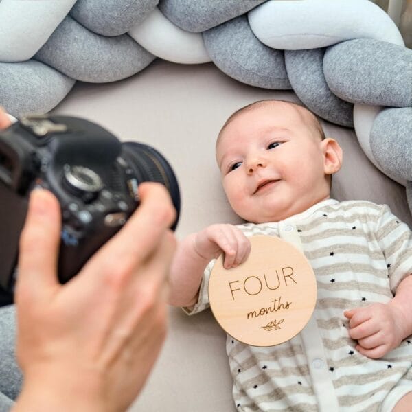 A baby lies on a bed holding a round wood piece that reads "Four months," perfect for capturing adorable newborn milestone photos as someone snaps a picture with a camera.
