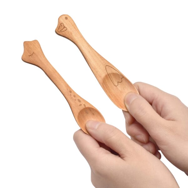 Hands holding two small, custom cartoon small wooden spoons. Wholesale wooden honey spoons available.