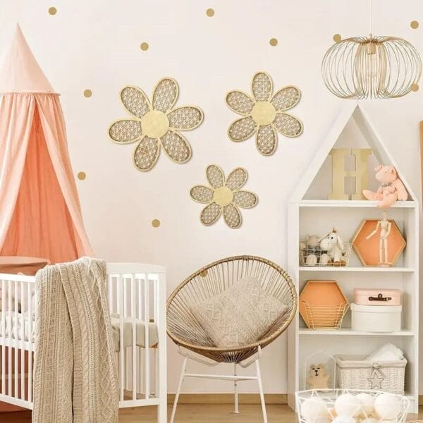 A nursery with a white crib, peach canopy, wicker chair, and house-shaped wall shelves; Bohemian wall decor accents like three floral hangings and gold polka dot decals add a playful touch to the space.