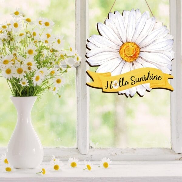 A white vase with daisies sits on a windowsill next to a "Hello Sunshine" wooden hanging plaque with a daisy and yellow ribbon. More daisies are scattered on the sill, enhancing this charming welcome sign display.