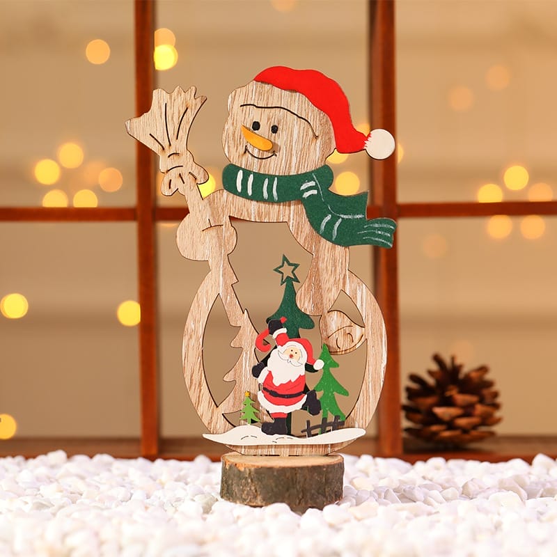 A Wood Christmas Decoration featuring a snowman with a Santa hat and green scarf stands on white pebbles, holding a broom, with a small Santa and Christmas tree at its base. Pine cone in the background. Custom Orders available.