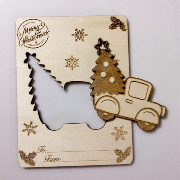A wooden Christmas card crafted from wood craft pieces, with cut-out shapes of a truck and tree, engraved snowflakes, "Merry Christmas" text, and "To/From" fields at the bottom.