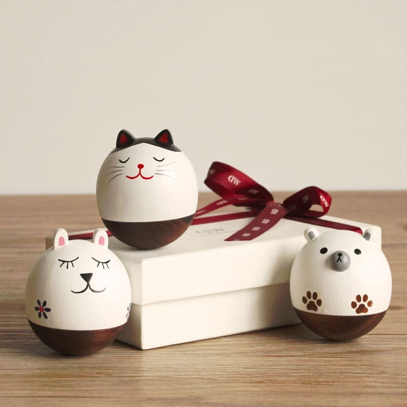 Three round animal figurines (cat, rabbit, bear), crafted as charming wooden crafts, are displayed on a wooden surface next to a white gift box with a red ribbon.