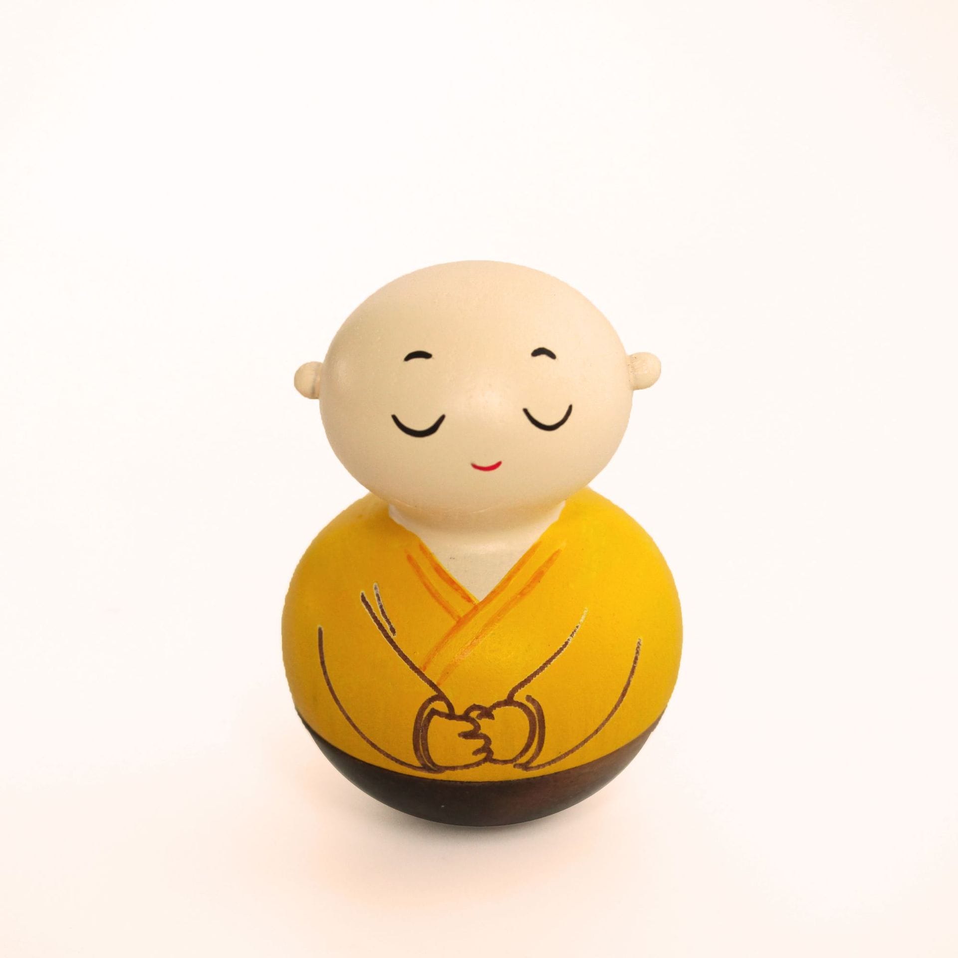A small, round wobbly toy figurine of a monk with closed eyes, a serene smile, and wearing a yellow robe, set against a plain white background.