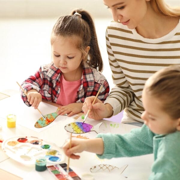 Two children and an adult paint vibrant fish drawings at a table, using watercolor paints and brushes to decorate Wood Fish Blanks that are colorable for kids.