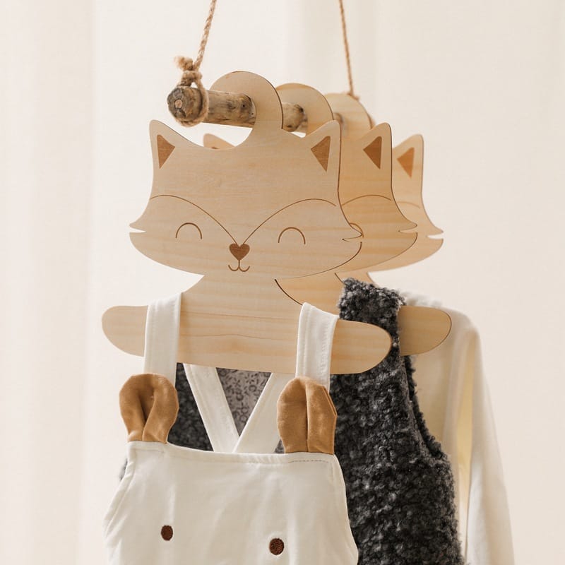 Three Nordic style baby hangers shaped like fox faces hold children’s clothing, including a fuzzy vest and overalls with animal ear details, hanging from a wooden rod.