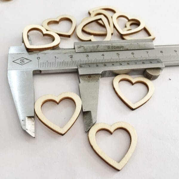 A caliper measures a wooden heart-shaped cutout, with several other eco-friendly wooden heart shapes scattered nearby on a white surface.