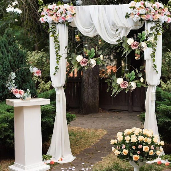 An outdoor wedding altar decorated with white fabric, pink and white flowers, greenery, and floral arrangements features wedding table decor and a bouquet of roses nearby for a charming celebration.