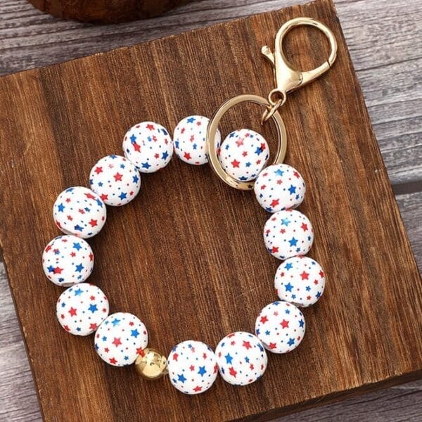 A beaded keychain bracelet with white beads decorated with red and blue stars, perfect for USA Independence Day, featuring a gold keyring and clasp, displayed on a wooden surface.