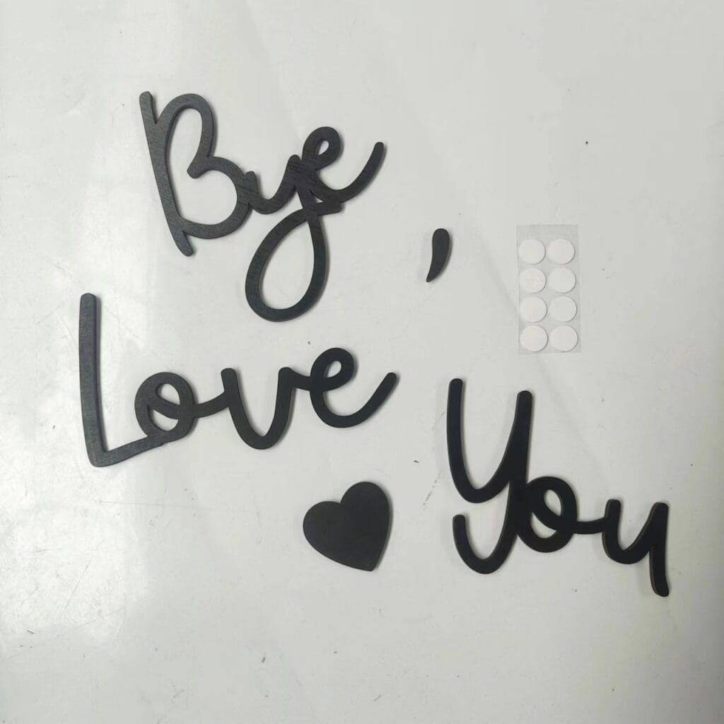 Black cutout words and wood letters spell "Bye, Love You" with a heart shape on a white surface; a small sheet of round adhesive stickers is nearby.