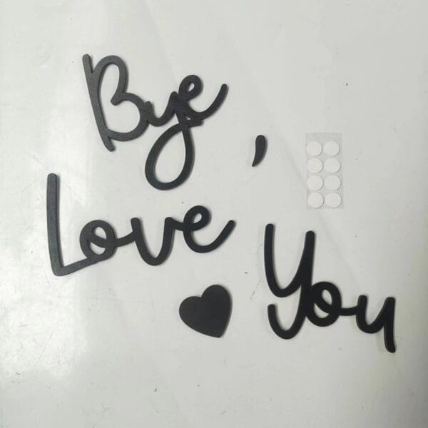 Black cutout words and wood letters spell "Bye, Love You" with a heart shape on a white surface; a small sheet of round adhesive stickers is nearby.