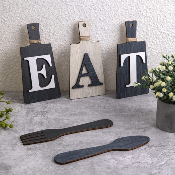 Three decorative wood sign boards with the letters E, A, and T stand upright; wooden fork and spoon cutouts are on the surface, next to small potted plants.