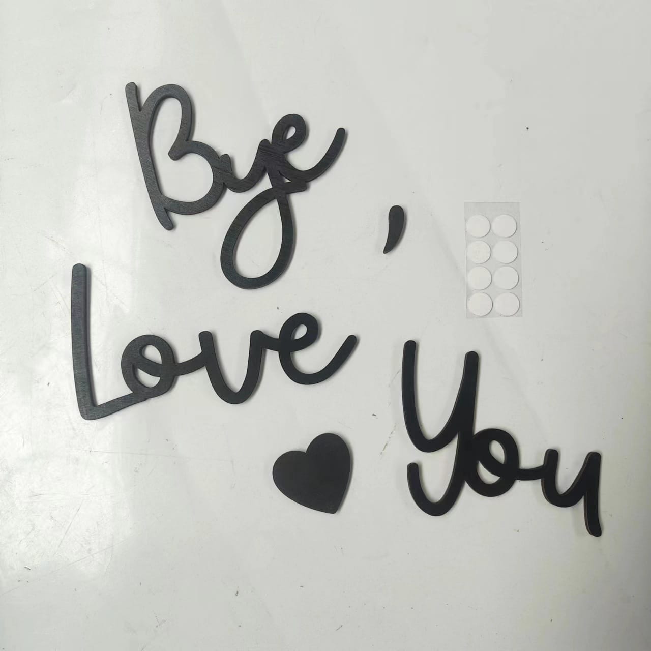 Black cut-out words "Bye, Love You" and a heart shape from a Letter Shape Set arranged on a white surface, with a small sheet of round adhesive tabs nearby—perfect for wood wall decor or wholesale craft projects.