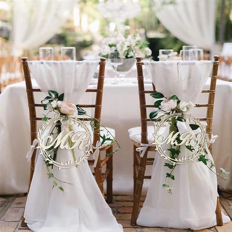 Two wooden chairs decorated with white fabric, greenery, and flowers, each with a sign reading "Mr." and "Mrs." in front of a table set for a wedding reception.