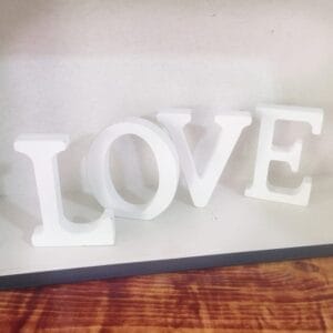 White woodcut letters spelling "LOVE" are arranged on a wooden surface against a light background, making a charming piece of home decor.