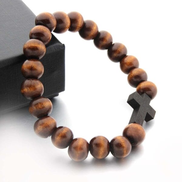 A wooden bead bracelet with a black wooden cross charm is displayed next to a sleek black box on a white surface.