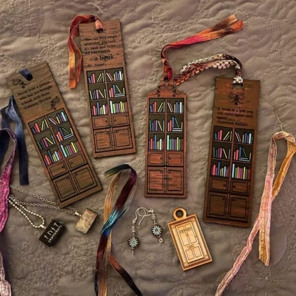Four illustrated wooden book mark designs feature bookshelf motifs, ribbons, and book-themed charms and jewelry, all artfully arranged on a textured gray quilt.