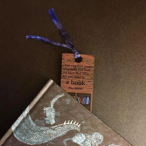 A brown wooden bookmark with a quote from Dr. Seuss is placed in a book featuring an illustrated dragon on its cover, both resting on a dark surface—perfect for bookmark craft enthusiasts or wholesale buyers.