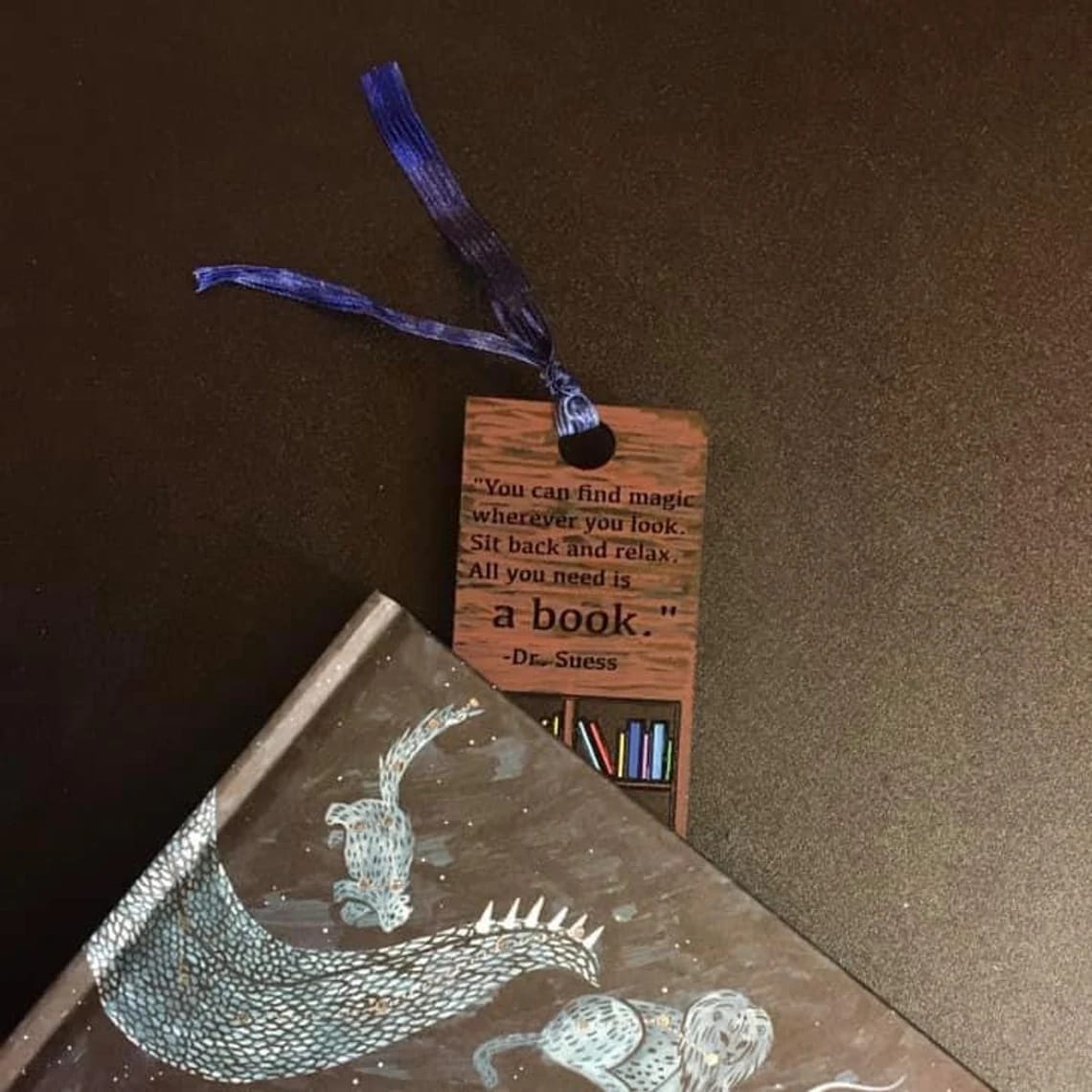 A brown wooden bookmark with a quote from Dr. Seuss is placed in a book featuring an illustrated dragon on its cover, both resting on a dark surface—perfect for bookmark craft enthusiasts or wholesale buyers.