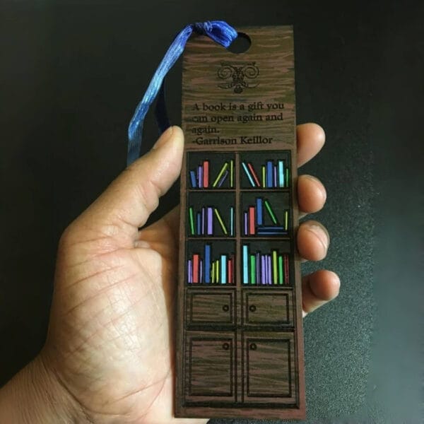A hand holds a wooden bookmark with a blue ribbon, featuring a bookshelf design and a Garrison Keillor quote—perfect for bookmark craft lovers or those seeking charming wholesale wooden bookmarks.