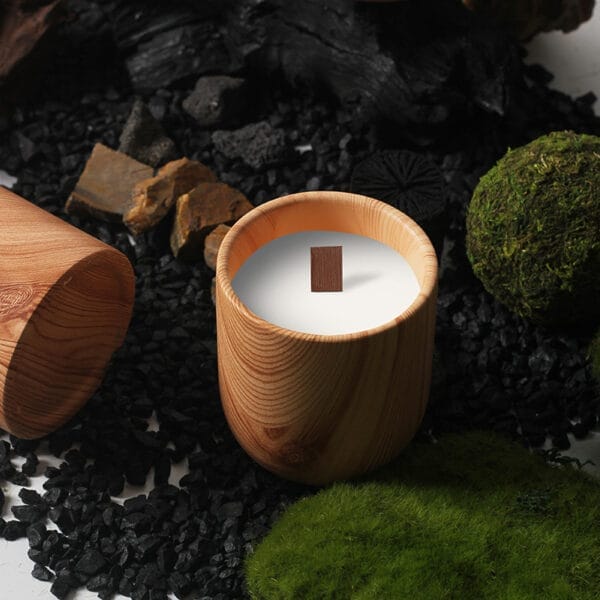 A Nordic minimalist candle holder with a white candle and wooden wick sits on a surface covered with black stones, moss, and rocks.